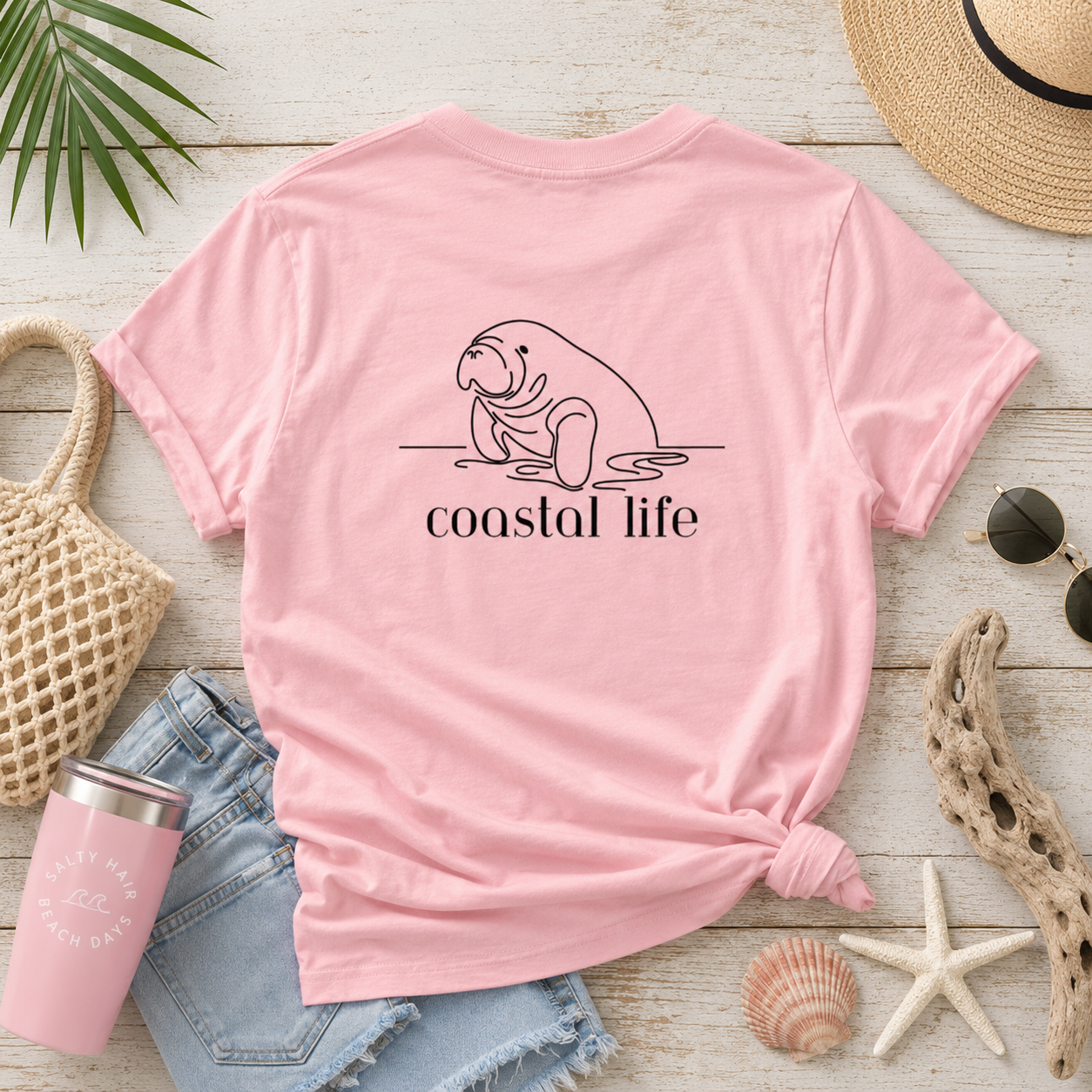 A pink short-sleeve T-shirt laid flat on a whitewashed wood surface, styled with beach-themed props including a straw hat, sunglasses, denim shorts, a pink tumbler, starfish, shells, and a woven bag. The shirt has a black line drawing of a manatee and the words 'coastal life' printed on the front.