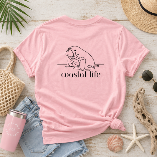 A pink short-sleeve T-shirt laid flat on a whitewashed wood surface, styled with beach-themed props including a straw hat, sunglasses, denim shorts, a pink tumbler, starfish, shells, and a woven bag. The shirt has a black line drawing of a manatee and the words 'coastal life' printed on the front.