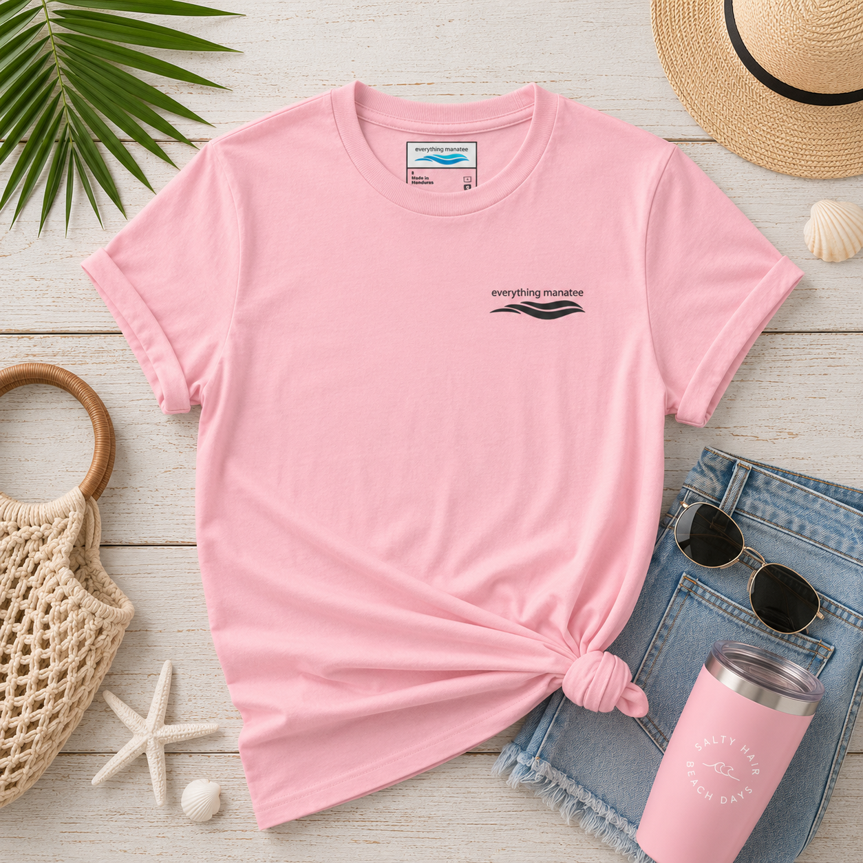 A pink short-sleeve t-shirt with a small black graphic on the left chest, styled in a flat lay with a knot at the hem. The shirt is surrounded by beach-themed props including a straw hat, denim shorts, sunglasses, a pink tumbler, a woven bag, and a palm leaf on a white wood background.