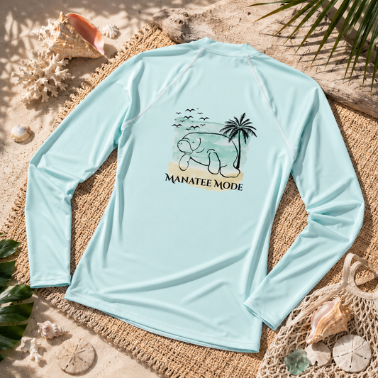 A light blue long-sleeve rash guard laid flat on a woven mat with beach-themed props including seashells, sand, and palm leaves. The shirt features a central graphic of a cartoon dog on a beach with a palm tree and the text 'MANATEE MODE'.