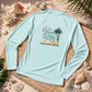 A light blue long-sleeve rash guard laid flat on a woven mat with beach-themed props including seashells, sand, and palm leaves. The shirt features a central graphic of a cartoon dog on a beach with a palm tree and the text 'MANATEE MODE'.