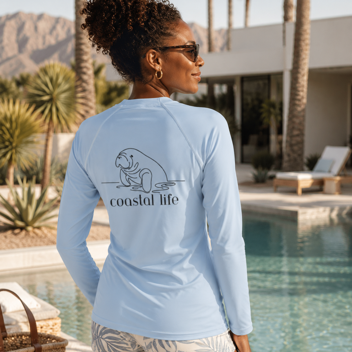 A woman stands by a swimming pool wearing a light blue long-sleeve rash guard, shown from the back. The shirt features a black line art graphic of a manatee and the text 'coastal life'.
