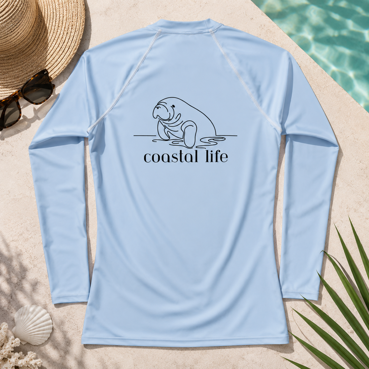 A light blue long-sleeve rash guard shirt laid flat on a sandy surface near a pool, with a straw hat, sunglasses, seashell, and palm leaves around it. The back of the shirt has a black line drawing of a manatee above the words 'coastal life'.