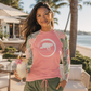 A woman wearing a pink long-sleeve rash guard with tropical palm leaf patterns on the sleeves and a white turtle logo on the chest, standing outdoors near a beach with palm trees and lounge chairs.