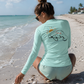A woman wearing a mint green long-sleeve rash guard and matching swim shorts is crouching on a sandy beach, viewed from behind to show the back graphic of a sleeping turtle with a sun and the word 'Turtle'.