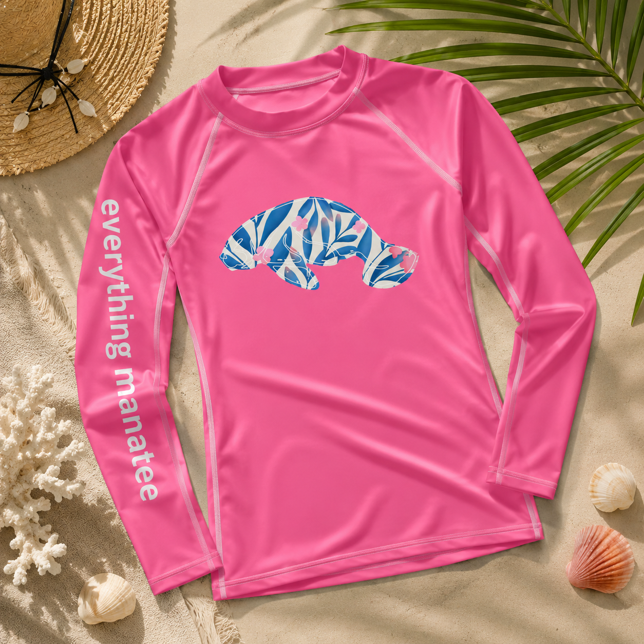 A bright pink long-sleeve rash guard laid flat on a sandy beach background with seashells, a straw hat, and palm leaves. The shirt has a blue and white tropical leaf patterned seal graphic on the chest and 'everything manatee' printed in white down one sleeve.