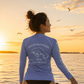 A woman wearing a light blue long-sleeve rash guard stands on a beach at sunset, facing away from the camera to show the back graphic that reads 'everything manatee ENJOY EVERY MOMENT'.