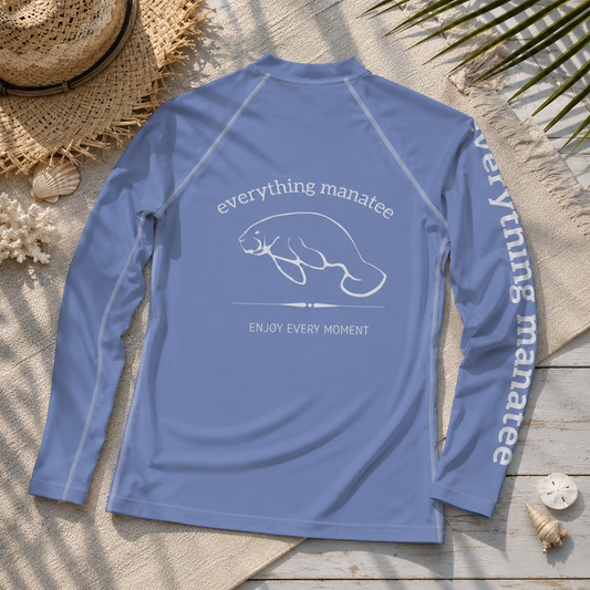 A light blue long-sleeve rash guard laid flat on a beach-themed background with a straw hat, seashells, and palm leaves. The back of the shirt features a white graphic of a manatee with the text 'everything manatee' and 'ENJOY EVERY MOMENT', and the left sleeve has 'everything manatee' printed vertically down the side.