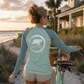 A woman stands on a beach path at sunset wearing a long-sleeve athletic shirt with a large white turtle logo on the back and the text 'Protect What You Love' near the hem. She is posed next to a vintage bicycle, with sand, beach grass, water, and a house in the background.