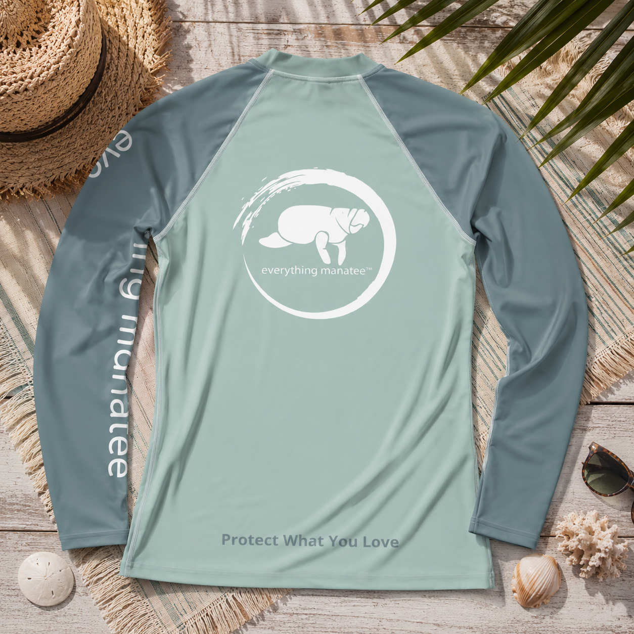 A long-sleeve raglan rash guard laid flat on a rustic wood surface with beach-themed props including a straw hat, palm leaves, shells, and sunglasses. The shirt is sage green with darker sage green sleeves and a white circular graphic on the chest featuring a turtle and the text 'everything marine'.