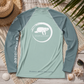 A long-sleeve raglan rash guard laid flat on a rustic wood surface with beach-themed props including a straw hat, palm leaves, shells, and sunglasses. The shirt is sage green with darker sage green sleeves and a white circular graphic on the chest featuring a turtle and the text 'everything marine'.