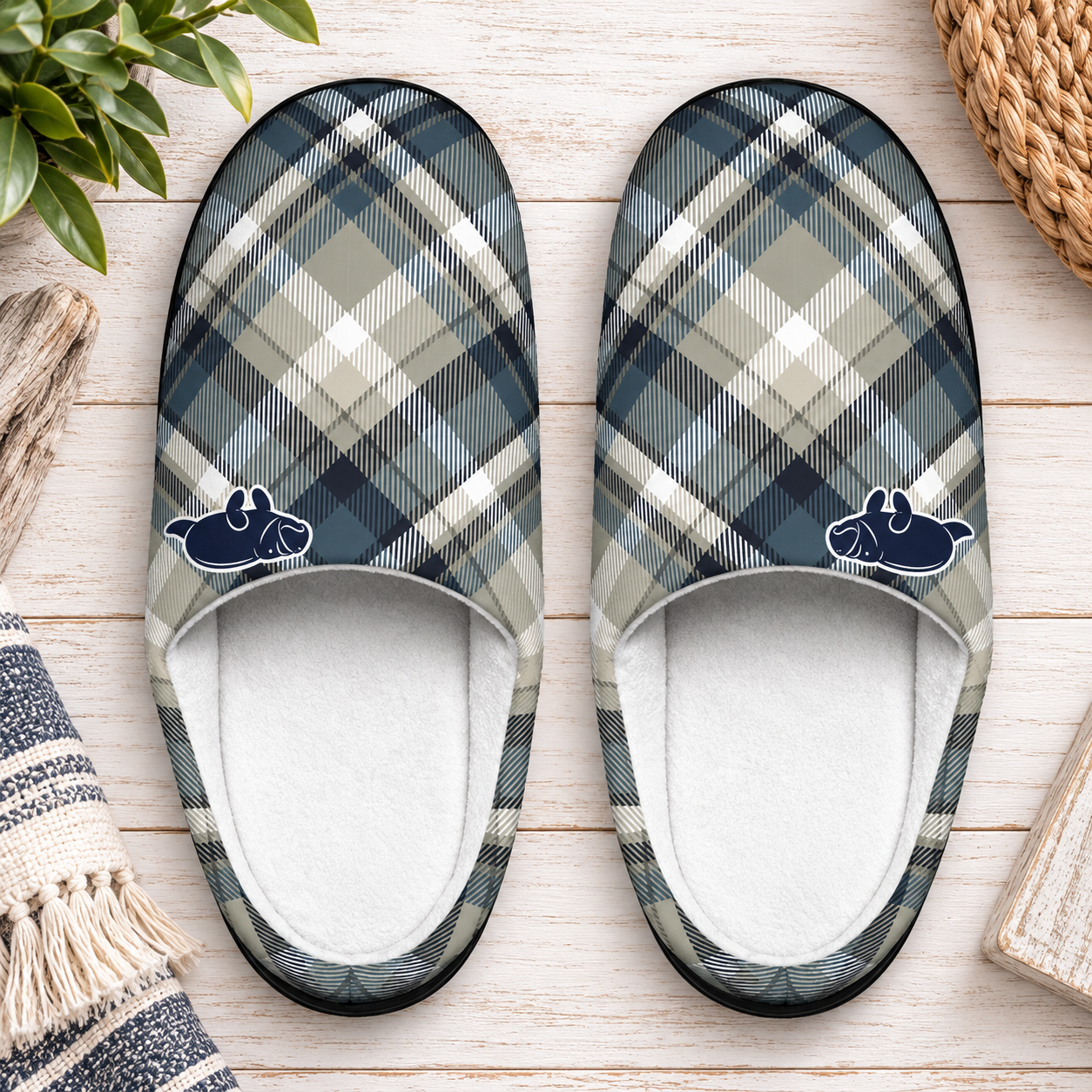 A pair of slip-on slippers with a blue, gray, and beige plaid pattern and small navy cow graphics near the opening, shown on a light wood surface with decorative props.