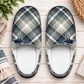 A pair of slip-on slippers with a blue, gray, and beige plaid pattern and small navy cow graphics near the opening, shown on a light wood surface with decorative props.