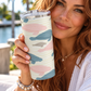 A woman holds a tall insulated tumbler with a clear lid. The tumbler has a cream background with abstract wavy stripes in dusty pink, slate blue, and light gray.