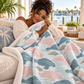 A woman sits on a beige sofa wrapped in a soft sherpa blanket featuring a dalmatian-style spot pattern in dusty pink, slate blue, and taupe on a light blue background. A tropical waterfront scene is visible in the background.