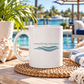 A white ceramic mug with the text 'everything manatee' and a teal wave graphic sits on a woven placemat by a poolside table. The background shows a tropical resort setting with palm trees, blue water, and lounge chairs.