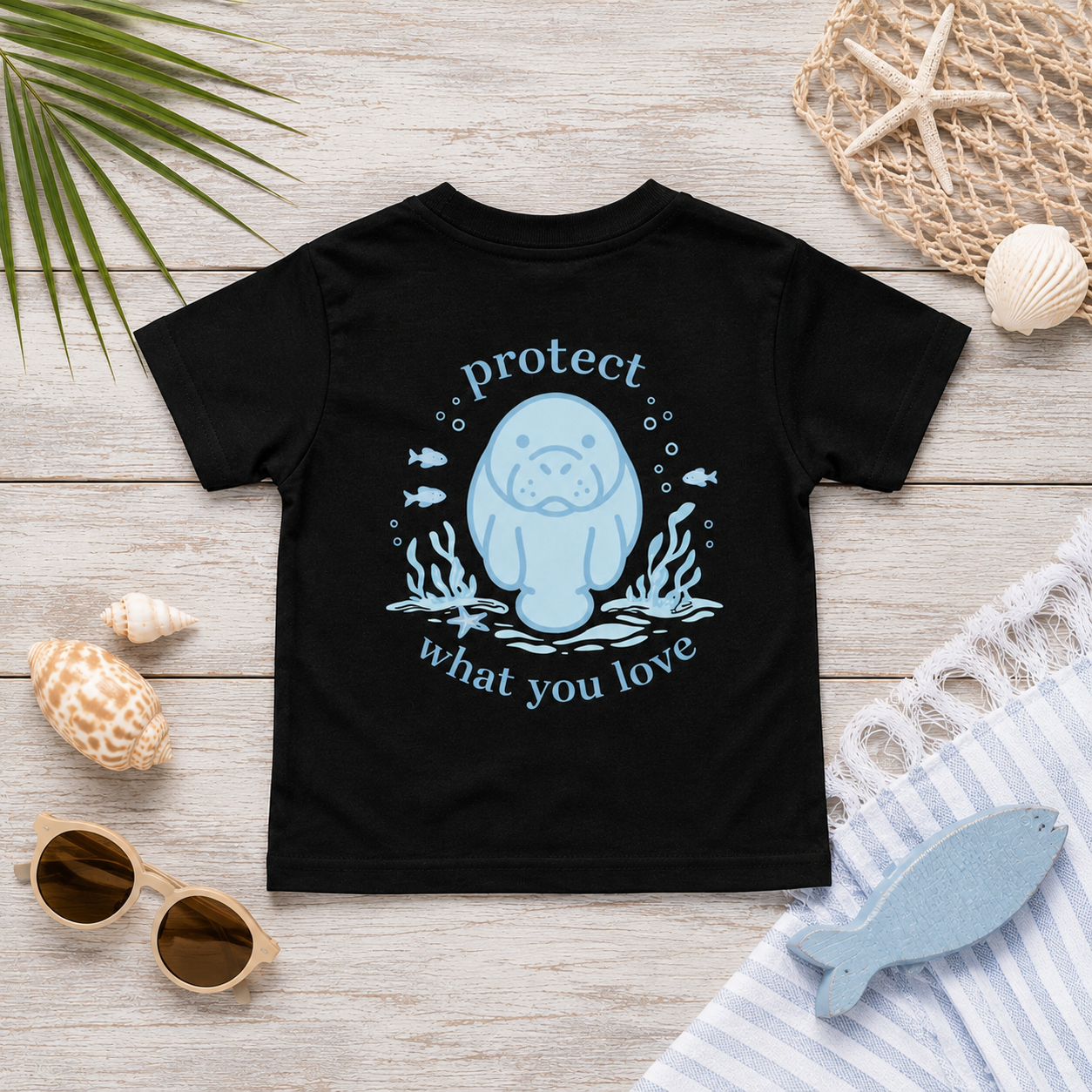 A black short-sleeve t-shirt with a light blue graphic of a manatee surrounded by the words 'protect what you love', styled in a flat-lay on a light wood background with beach-themed props.