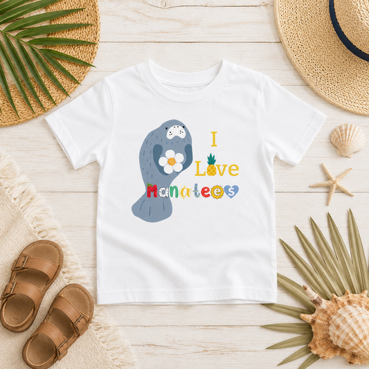 A white kids' t-shirt with a colorful graphic of a manatee holding a daisy and the text 'I Love Hanape'eos', styled in a flat-lay with beach-themed props like sandals, a straw hat, and seashells on a light wood background.
