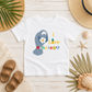 A white kids' t-shirt with a colorful graphic of a manatee holding a daisy and the text 'I Love Hanape'eos', styled in a flat-lay with beach-themed props like sandals, a straw hat, and seashells on a light wood background.