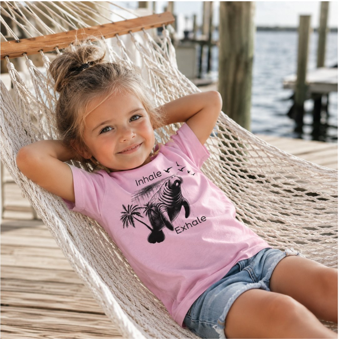 A young girl with her hair in a bun relaxes in a woven hammock on a wooden dock by the water. She is wearing a light pink graphic t-shirt with a black seal print and the words 'Inhale.