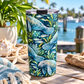 A tall insulated tumbler with a lid sits on a wooden table outdoors near the water, decorated with a blue and green manatee and sea plant pattern. A starfish, woven mat, plant, and driftwood are visible around it.