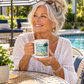 A smiling older woman sits at an outdoor patio table by a pool, holding a white ceramic mug with a teal and black tropical graphic and the words “MANATEE MODE.” She wears a white crochet top and sunglasses rest on her head.