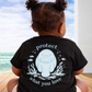 Child wearing a black t-shirt with an environmental message and cartoon character, sitting by a window with a view of the ocean.