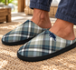 A close-up of a person wearing plaid slip-on slippers with a navy blue mascot logo on the side, standing on a woven rug in a sunlit indoor setting.