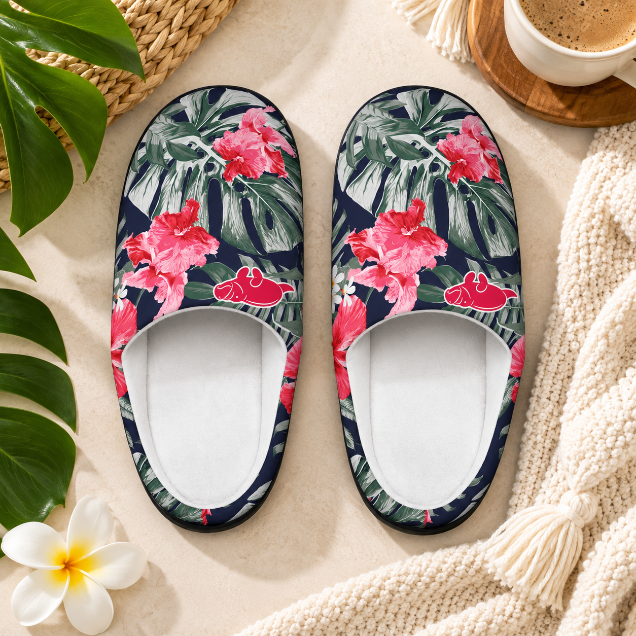A pair of slip-on house slippers with a tropical floral print featuring pink hibiscus flowers and green leaves on a dark navy background, styled on a beige surface with a knit blanket, a cup of coffee, and green leaves.