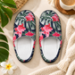 A pair of slip-on house slippers with a tropical floral print featuring pink hibiscus flowers and green leaves on a dark navy background, styled on a beige surface with a knit blanket, a cup of coffee, and green leaves.