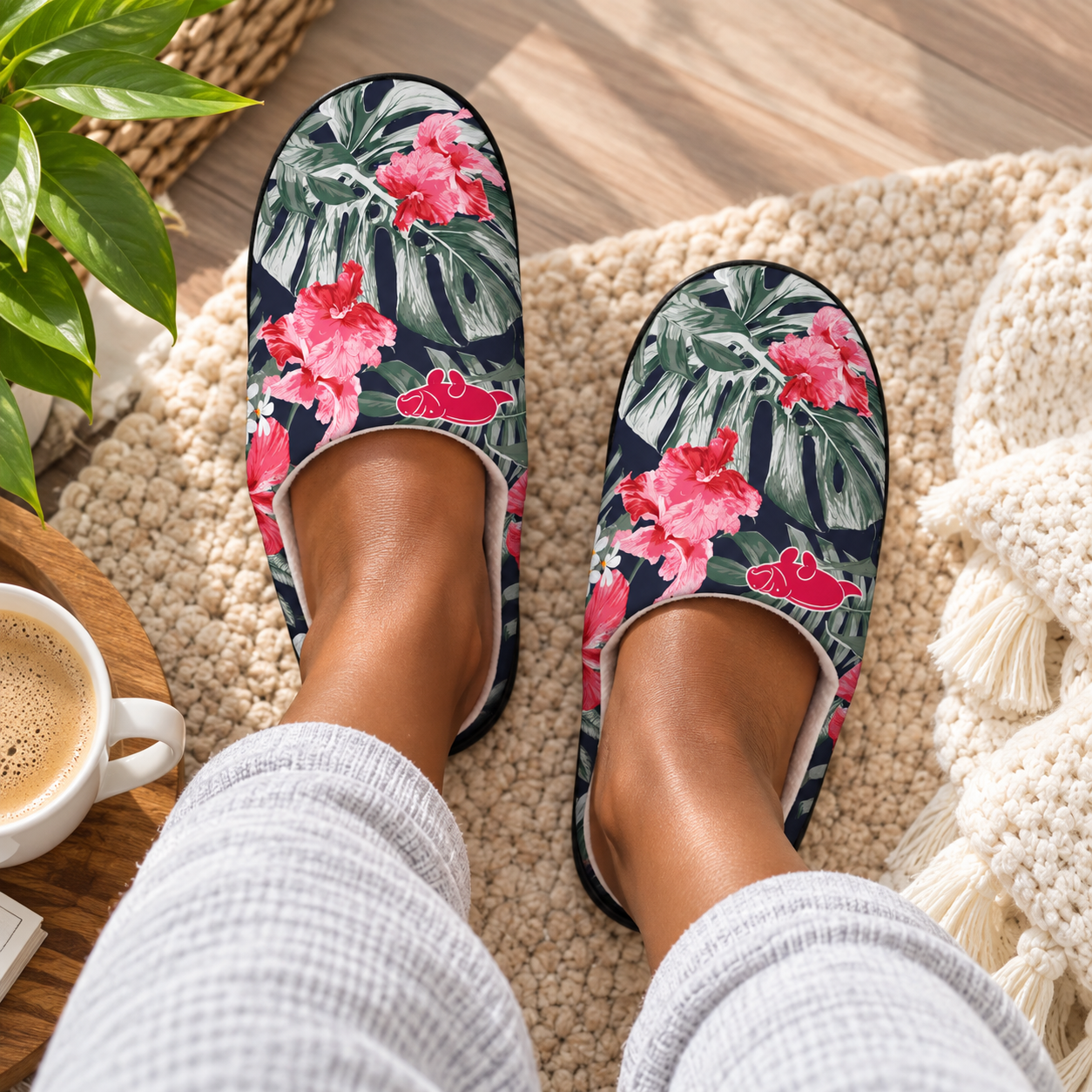A person wearing a pair of slip-on slippers with a tropical floral print featuring pink flowers and green leaves on a dark navy background, sitting on a beige knit rug with a coffee cup and plant nearby.