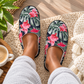 A person wearing a pair of slip-on slippers with a tropical floral print featuring pink flowers and green leaves on a dark navy background, sitting on a beige knit rug with a coffee cup and plant nearby.