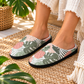 A person wearing tropical-print slip-on slippers with a black sole, standing on a woven straw rug near a potted plant.