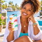 A smiling woman with curly hair sits by the water holding a white insulated tumbler with colorful turtle illustrations. She wears a turquoise top, white button-up shirt, gold necklace, and beaded bracelets.