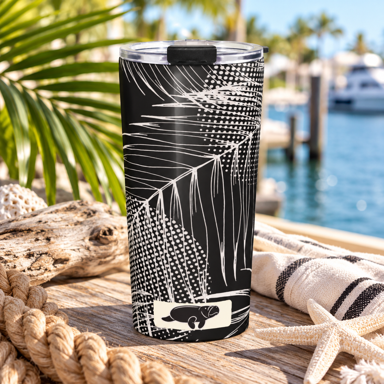 A black insulated tumbler with a white tropical palm leaf pattern and a small white logo near the bottom, shown on a wooden dock with water, boats, palm leaves, rope, and a striped towel in the background.