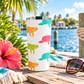 A white insulated tumbler with a lid sits on a wooden dock by the water, decorated with colorful manatee silhouettes. Tropical plants, a pink hibiscus flower, sunglasses, and a blue-and-white fringed towel are around it, with a sunny outdoor waterfront scene in the background.
