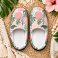 A pair of slip-on house slippers with a tropical print featuring pink pineapples, green palm leaves, and black silhouettes, displayed on a woven straw mat with decorative tropical leaves and a hibiscus flower.