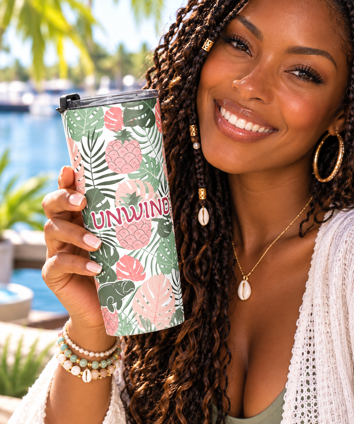 A smiling woman holds a insulated tumbler with a tropical leaf and pineapple pattern in green, pink, and white, featuring the word 'UNWINE' in pink. The background shows a blurred outdoor poolside setting with palm trees.