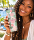 A smiling woman holds a insulated tumbler with a tropical leaf and pineapple pattern in green, pink, and white, featuring the word 'UNWINE' in pink. The background shows a blurred outdoor poolside setting with palm trees.