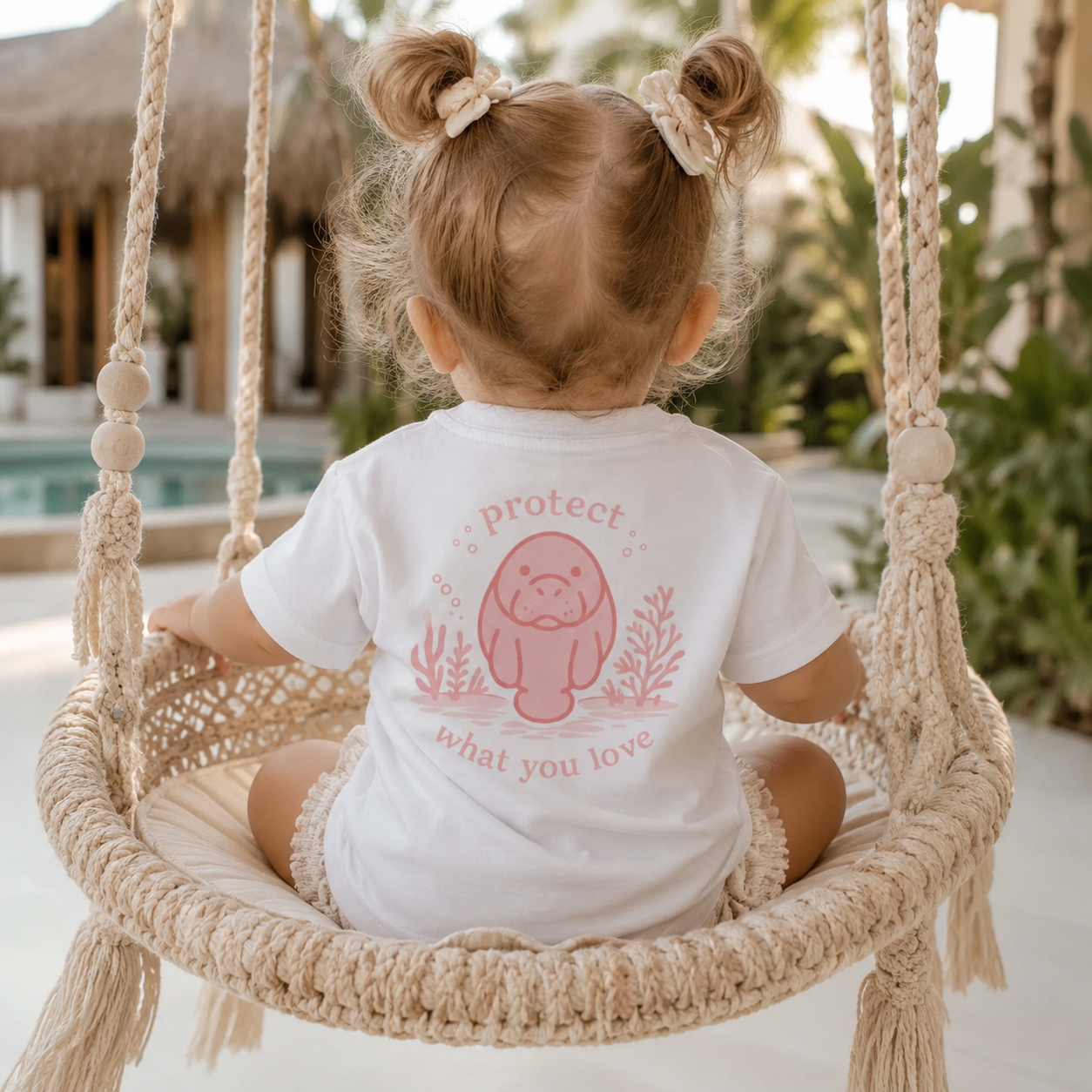 A toddler seen from behind sitting in a woven macrame swing, wearing a white short-sleeve bodysuit with a pink cartoon seal graphic and the words 'protect what you love' on the back. The setting is a bright, sunny patio with a pool and tropical plants in the background.