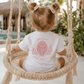 A toddler seen from behind sitting in a woven macrame swing, wearing a white short-sleeve bodysuit with a pink cartoon seal graphic and the words 'protect what you love' on the back. The setting is a bright, sunny patio with a pool and tropical plants in the background.