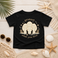 A black short-sleeve t-shirt with a beige graphic of three manatees and the text 'protect what you love', styled in a flat lay on a white wood background with beach-themed props like starfish, shells, netting, and sunglasses.