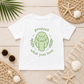 A white short-sleeve kids' T-shirt laid flat on a light wood surface, featuring a green cartoon manatee graphic with the words 'protect what you love.' The shirt is styled with beach-themed props including seashells, a starfish, woven mats, sunglasses, and a palm leaf.