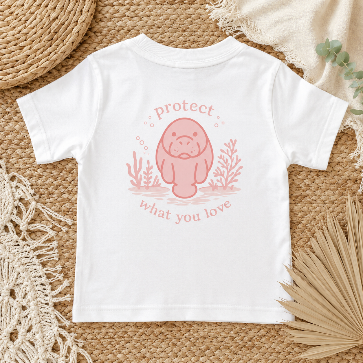 A white short-sleeve baby t-shirt with a pink cartoon manatee graphic and the words 'protect what you love' printed on the back, styled flat on a woven straw mat with boho props.