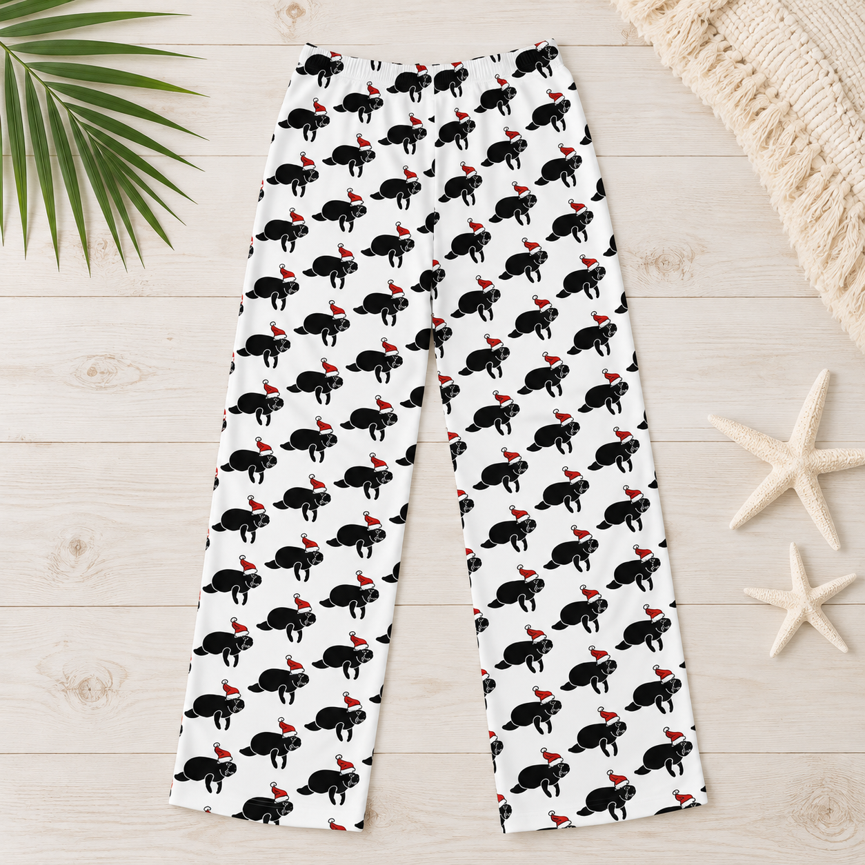 White wide-leg pants with an all-over black cat print wearing red Santa hats, laid flat on a light wood surface with palm leaf, starfish, and woven textile props.