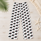 White wide-leg pants with an all-over black cat print wearing red Santa hats, laid flat on a light wood surface with palm leaf, starfish, and woven textile props.