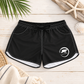 Black swim shorts with white contrast stitching, a black drawstring waist, and a white circular logo on the lower left leg, displayed in a flat-lay arrangement on a light wood background with palm leaves, driftwood, a seashell, and a woven towel.