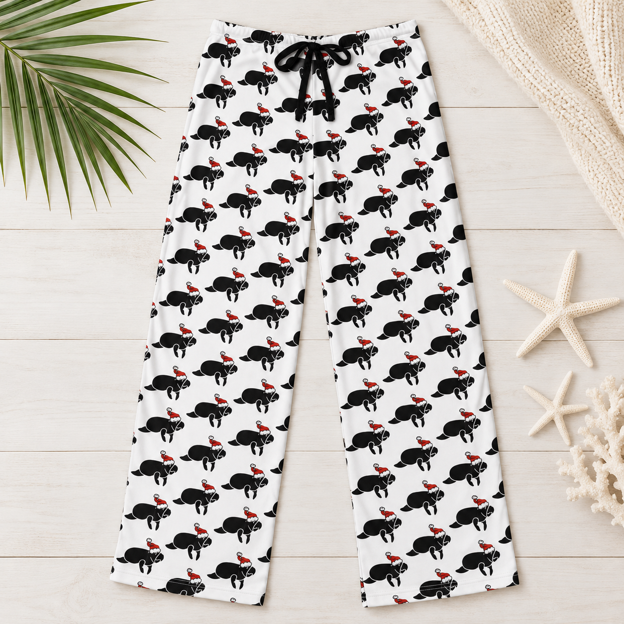 White lounge pants with an all-over black cat print wearing red Santa hats, styled in a flat lay on a light wood background with palm leaves, a knit blanket, and starfish.