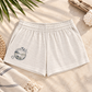 A pair of light beige heathered shorts with an elastic waistband and a circular 'Cape Town Coast' graphic on the left leg, styled in a beach-themed flat lay with palm leaves, a woven basket, and a striped towel.