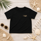 A black short-sleeve t-shirt with a small gold 'everything is okay' graphic on the left chest, styled in a flat lay on a light wood background with beach-themed props including seashells, a starfish, sunglasses, and woven mats.